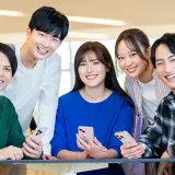 スマートフォンを持つ学生が集まって笑顔で話している大学生